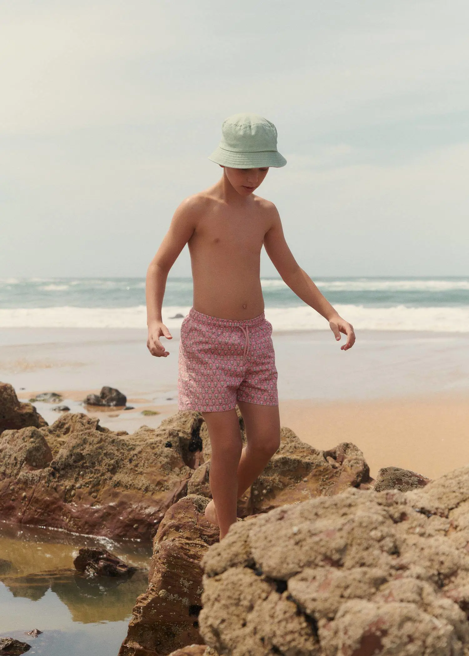 Mango KIDS/ Floral print swimsuit. a young man walking on a rock on the beach. 