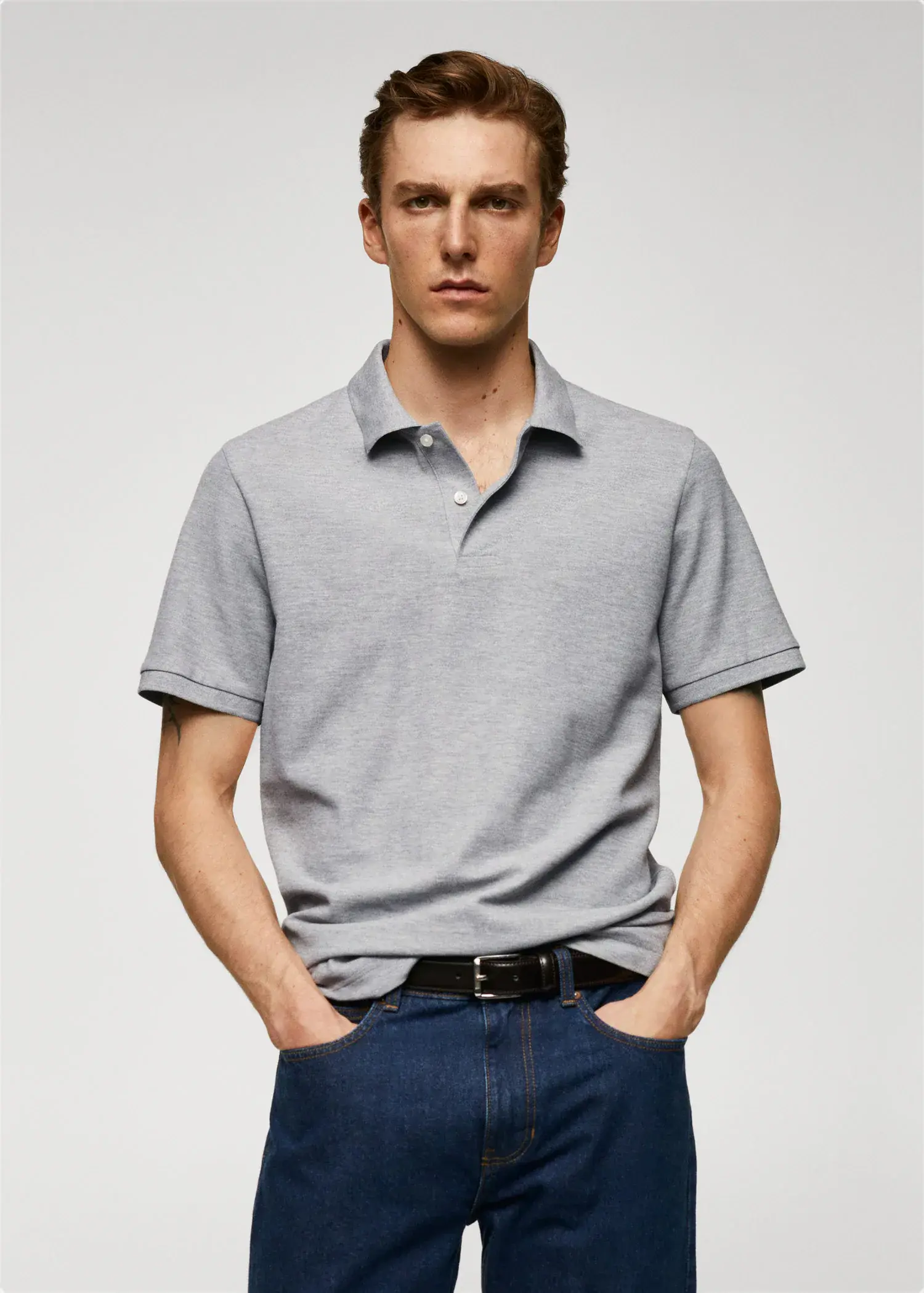 Mango 100% cotton pique polo shirt. a man in a gray polo shirt is standing with his hands in his pockets 