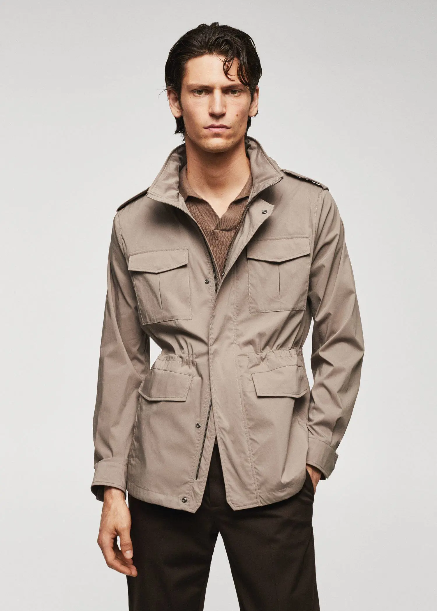 Mango Lightweight saharian jacket with pockets. a man in a tan jacket standing in front of a white wall. 