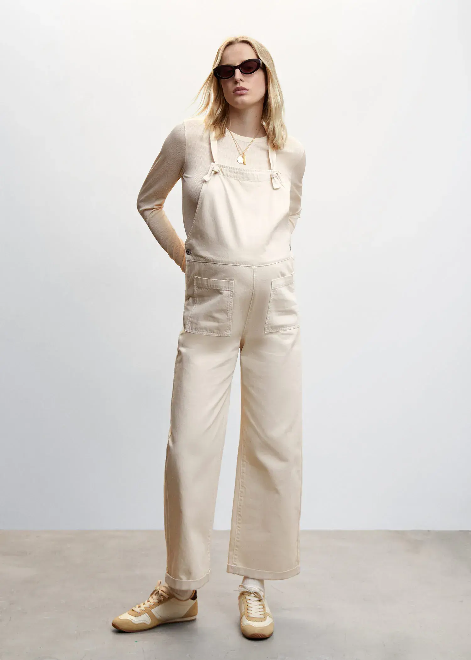 Mango Maternity denim dungarees. a woman wearing a white jumpsuit standing in front of a wall. 