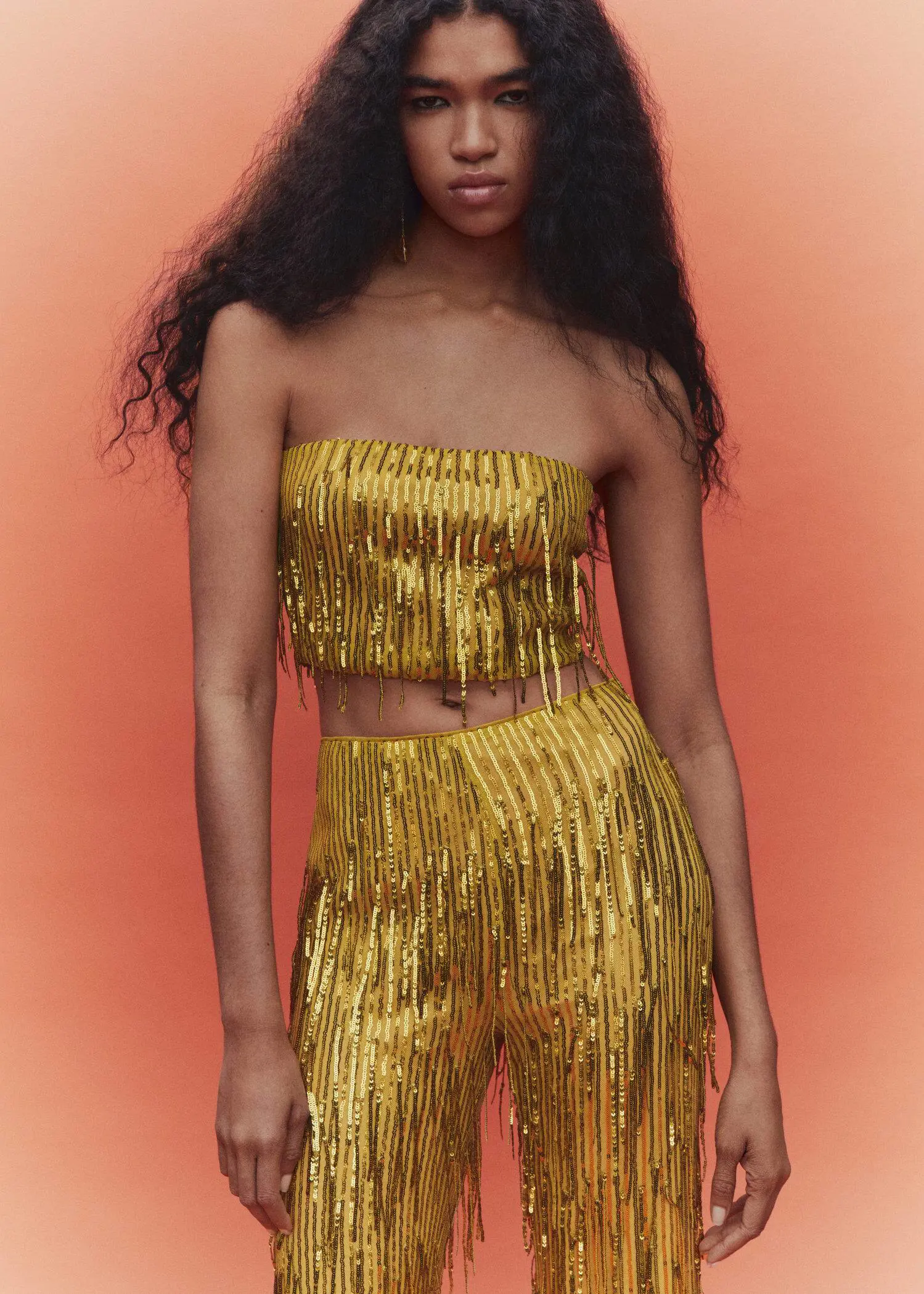 Mango Metallic fringed top. a woman in a gold outfit standing in front of an orange background. 