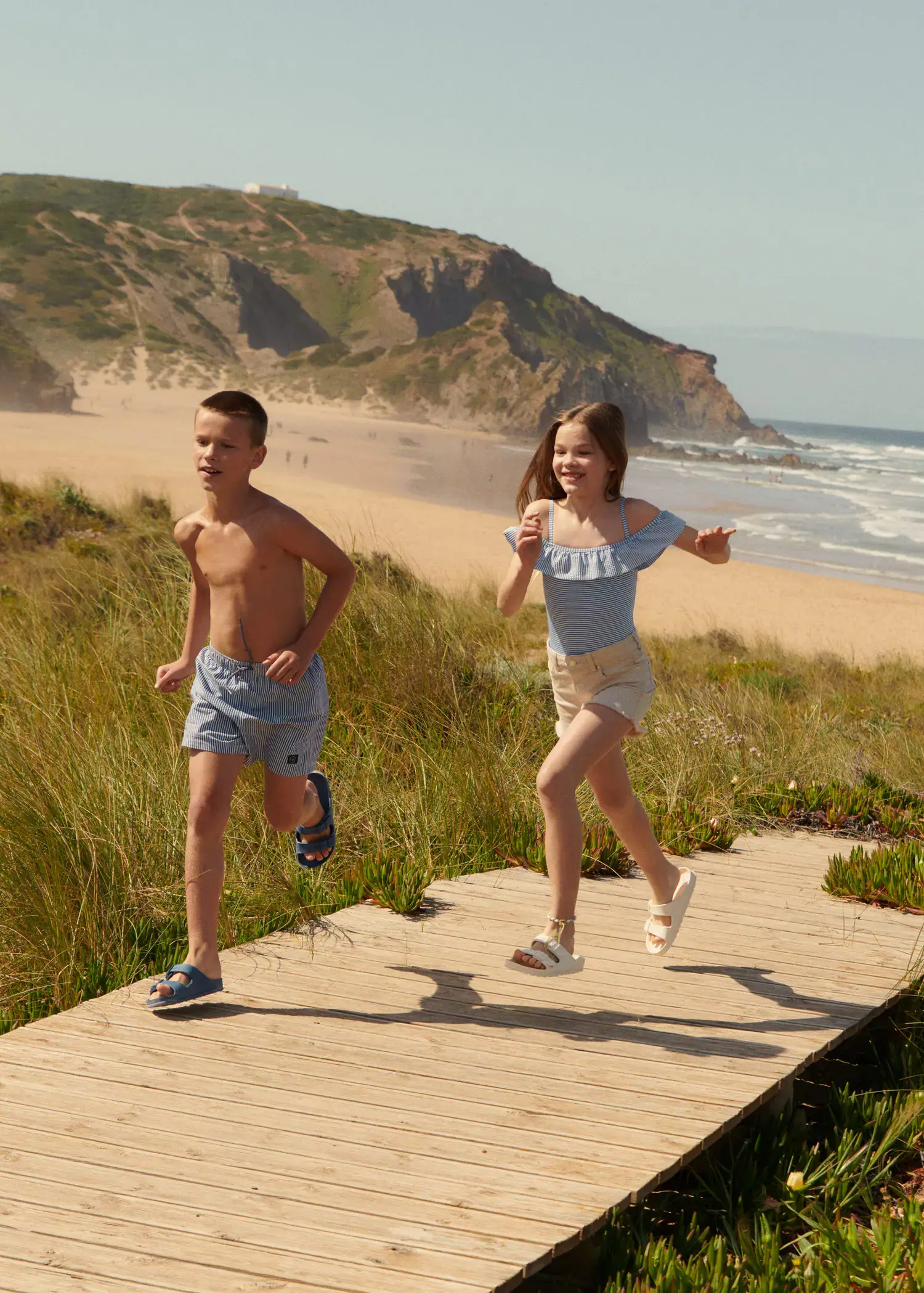 Mango KIDS/ Striped swimsuit. two children running on a boardwalk near the beach. 