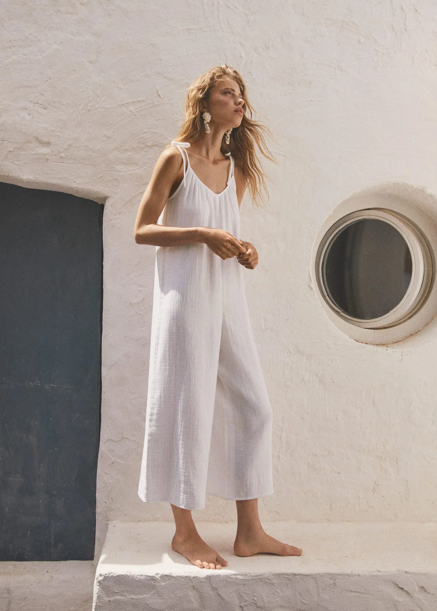 Mango Textured jumpsuit with bows. a woman in a white dress standing in front of a window. 