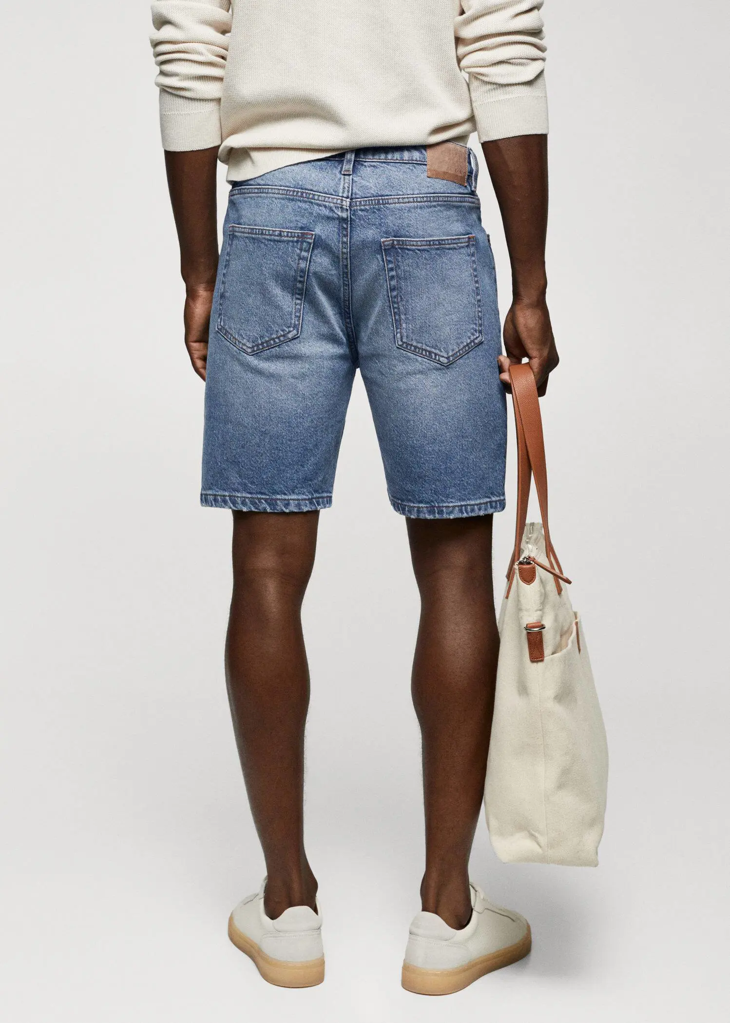 Mango Regular-fit denim bermuda shorts. a man holding a white bag in front of a wall. 